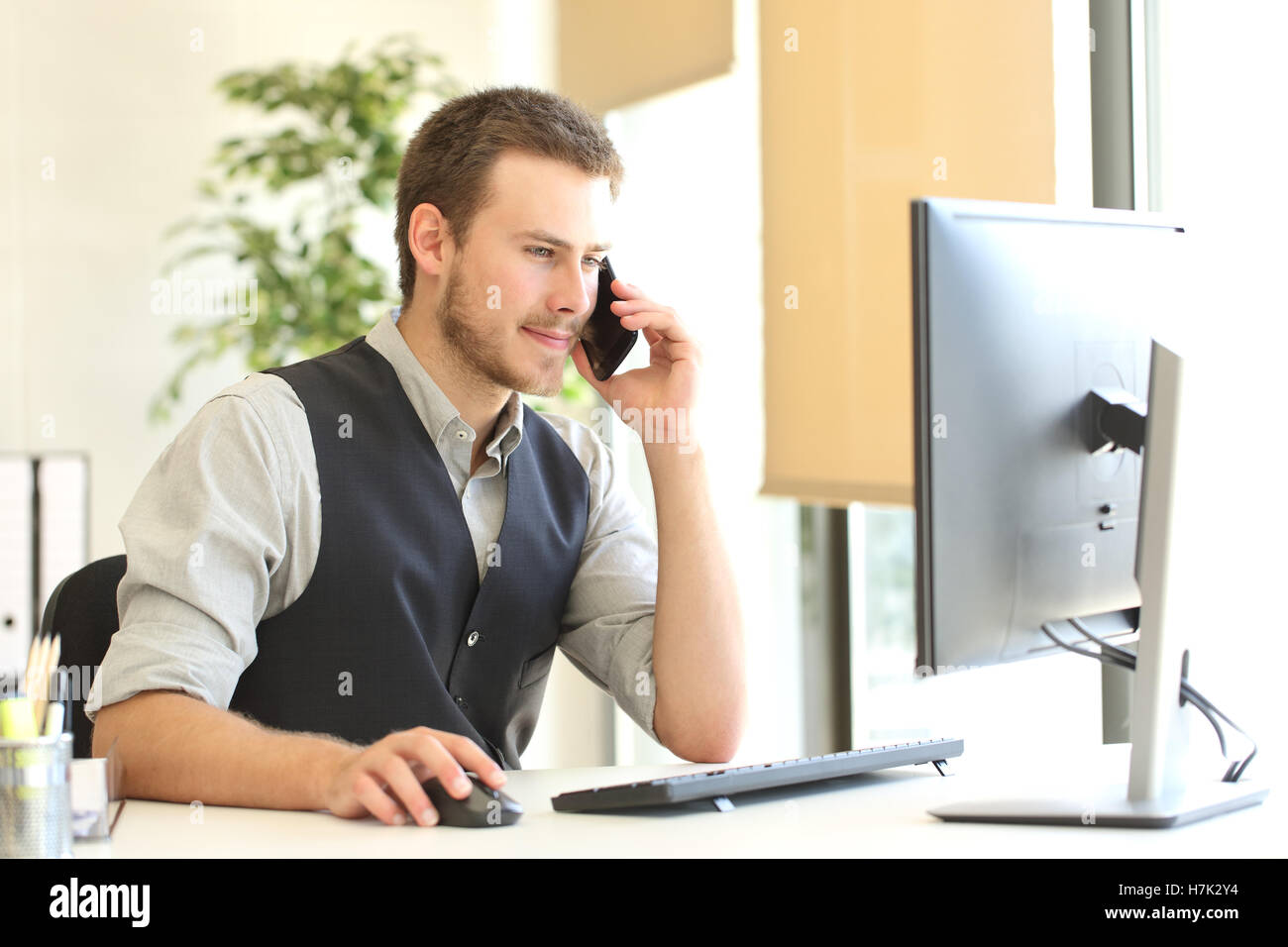 Imprenditore chiamando sul telefono per il servizio di assistenza al cliente e utilizzando un computer desktop in ufficio con una finestra in background Foto Stock