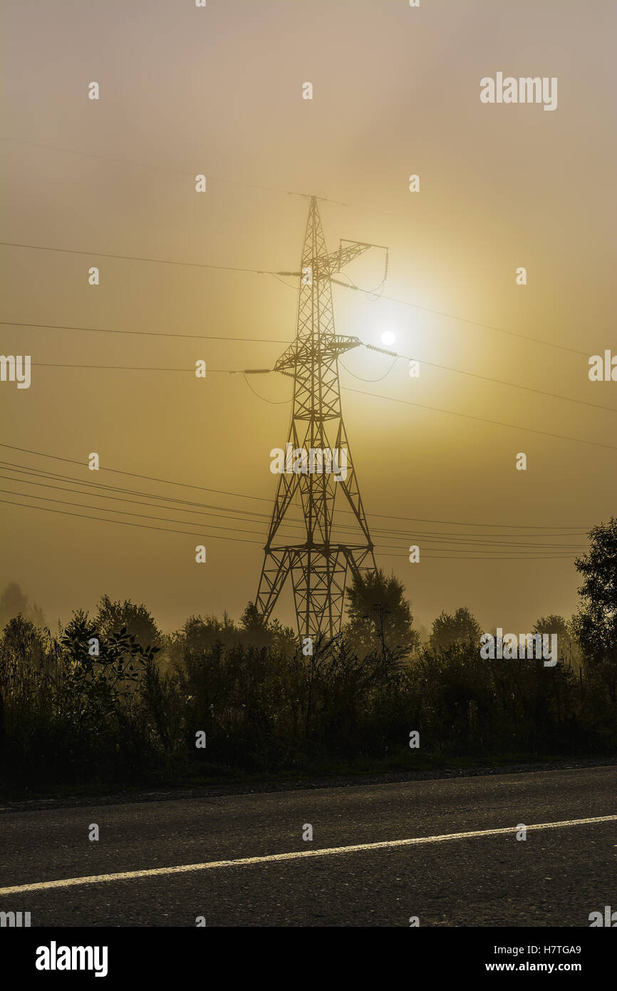 Alta tensione La torre di trasmissione vicino strada rurale. Energia elettrica sul filo di nebbia di mattina sfondo. Foto Stock