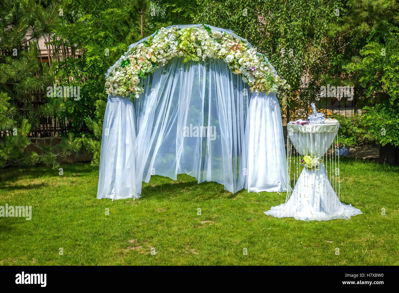 Decorazioni per matrimoni. Arco fiorito e tabella coperta di chiffon bianco preparato per la cerimonia di matrimonio nel verde giardino. Foto Stock