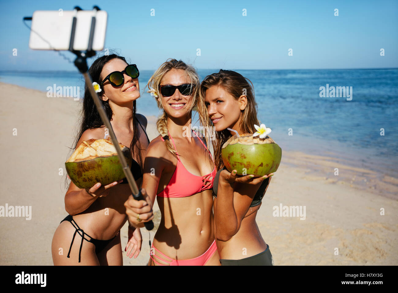 Gruppo di giovane donna azienda noci di cocco e tenendo selfie sulla spiaggia. Tre giovani donne in costume da bagno sulla spiaggia prendendo self por Foto Stock