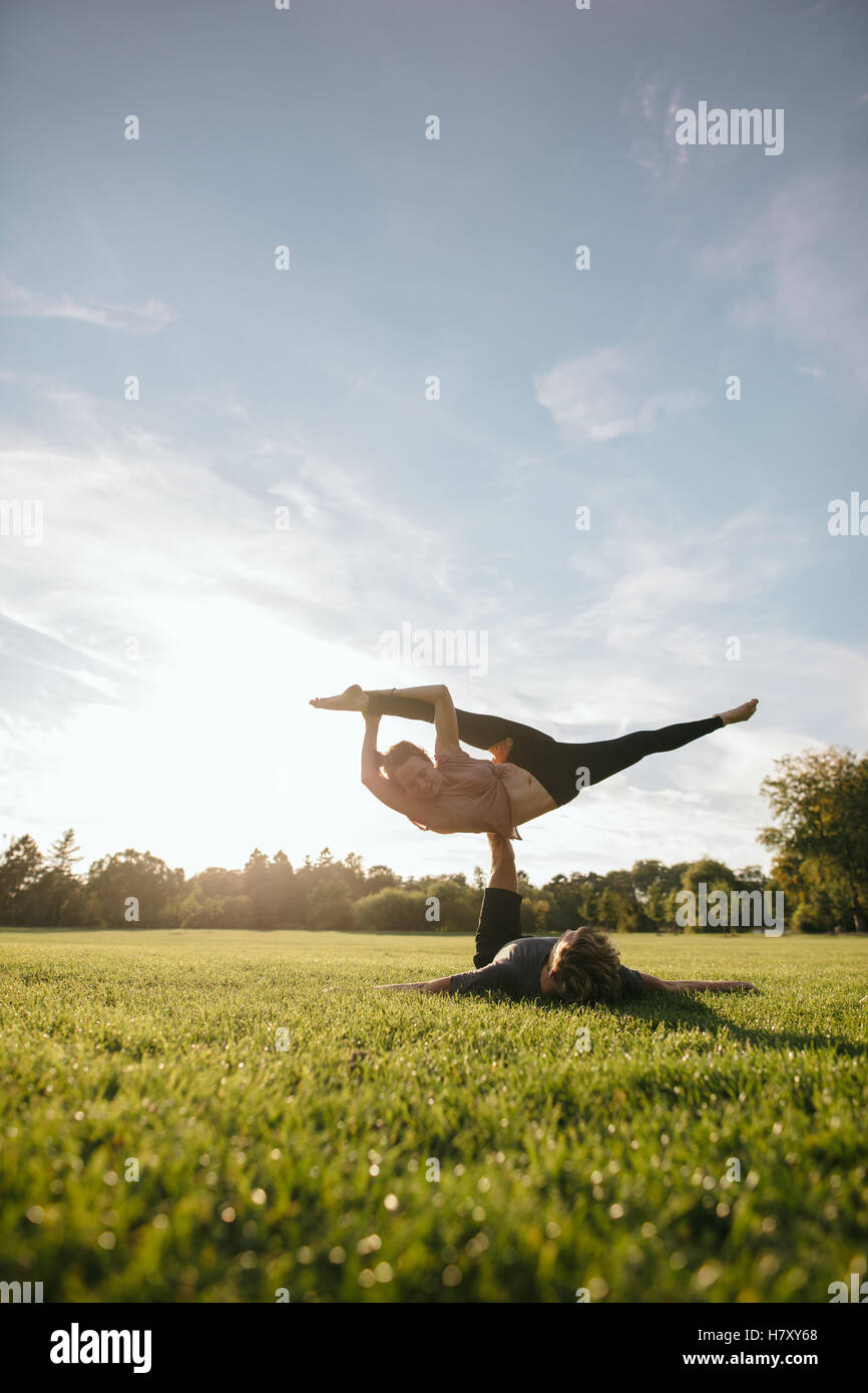 Colpo verticale di adattamento coppia giovane facendo yoga acrobatico. Uomo disteso sull'erba e il bilanciamento della donna in piedi presso il parco. Foto Stock