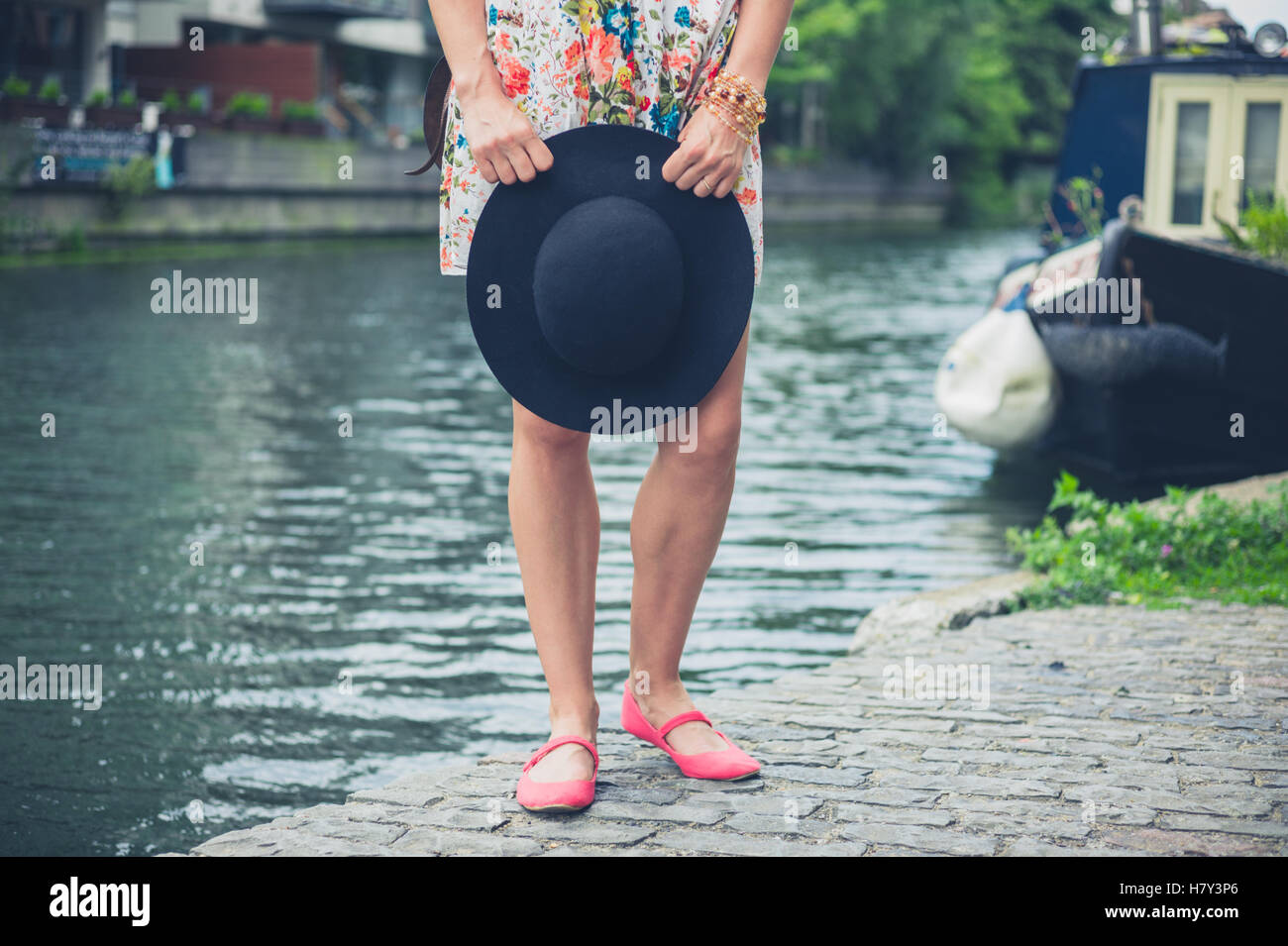 Una giovane donna che indossa un abito e tenendo un cappello è in piedi da un canale con una casa galleggiante in background Foto Stock