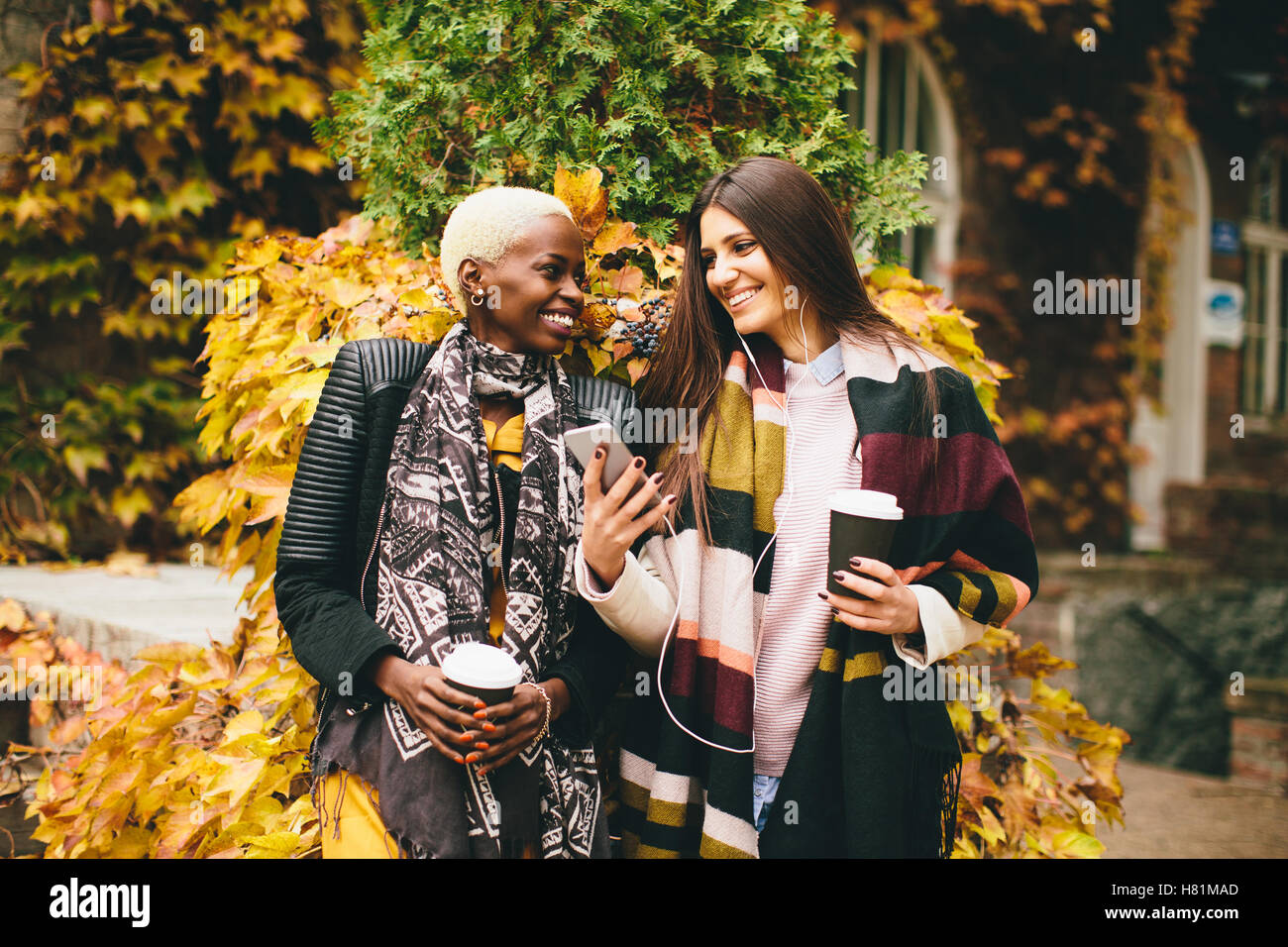 Americano africano e caucasici che pongono la donna fuori con il telefono cellulare e una tazza di caffè per andare in autunno Foto Stock