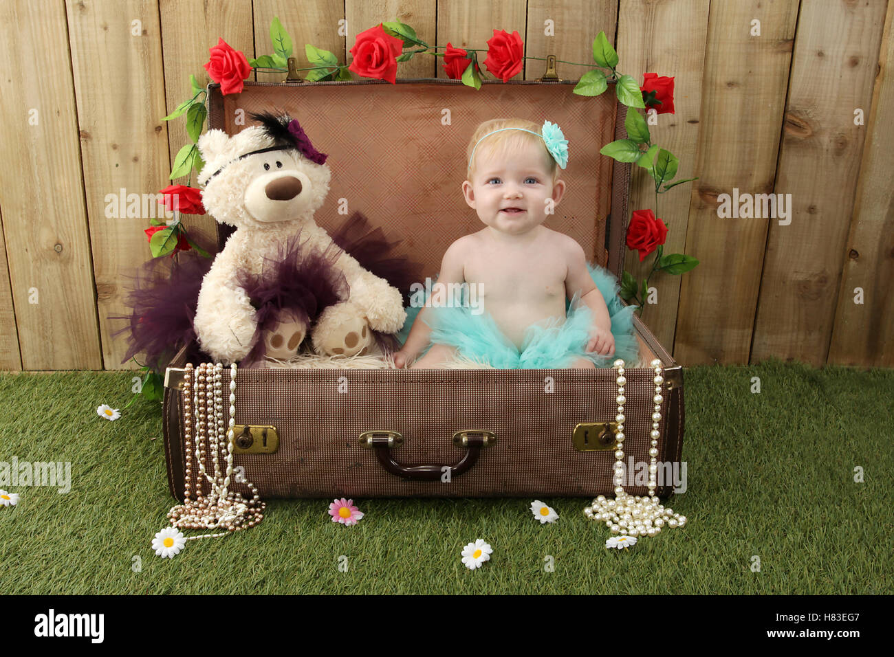 Baby ragazza seduta in una valigia in giardino con erba e fiori Foto Stock