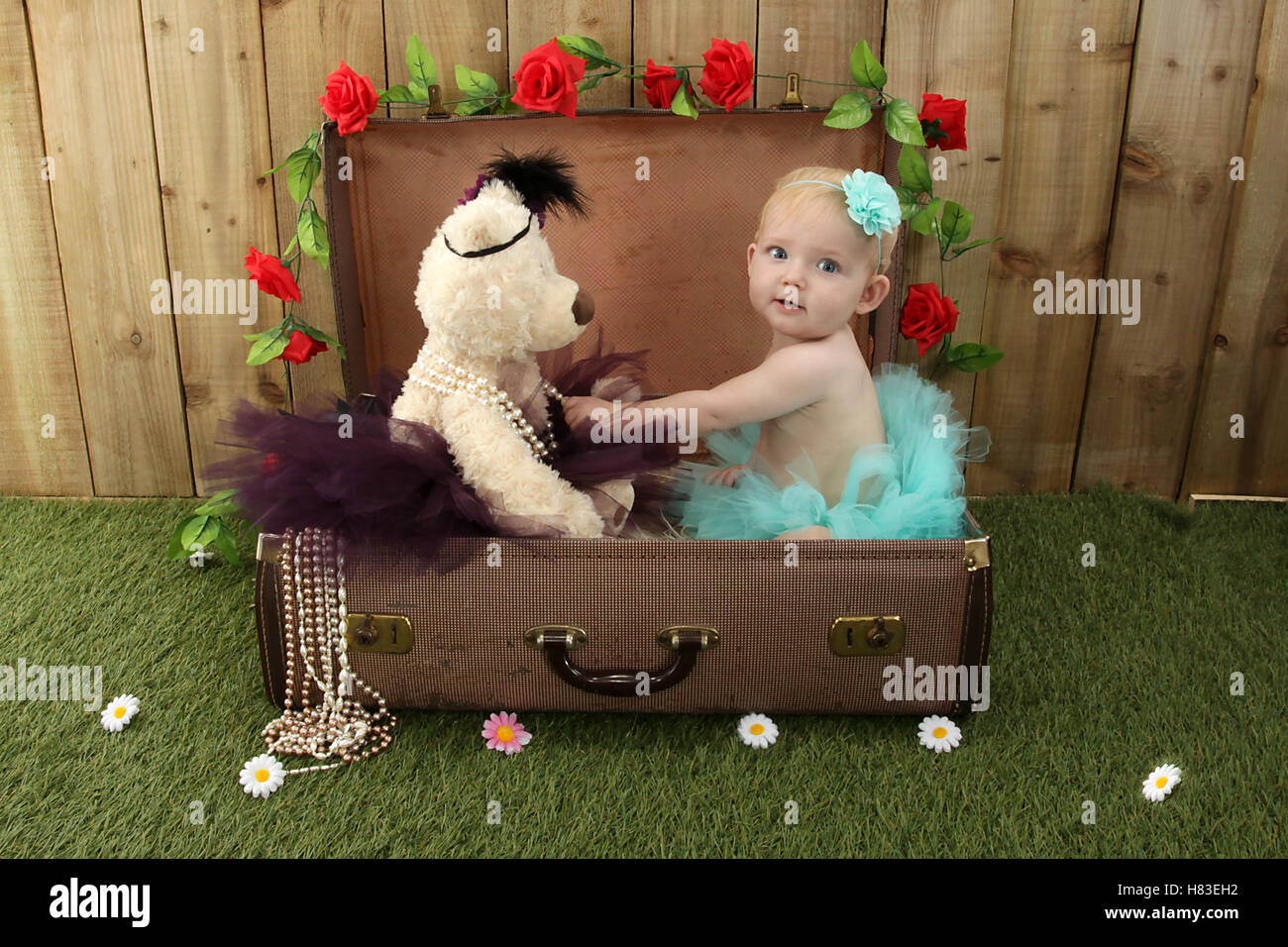 Baby ragazza seduta in una valigia in giardino con erba e fiori Foto Stock