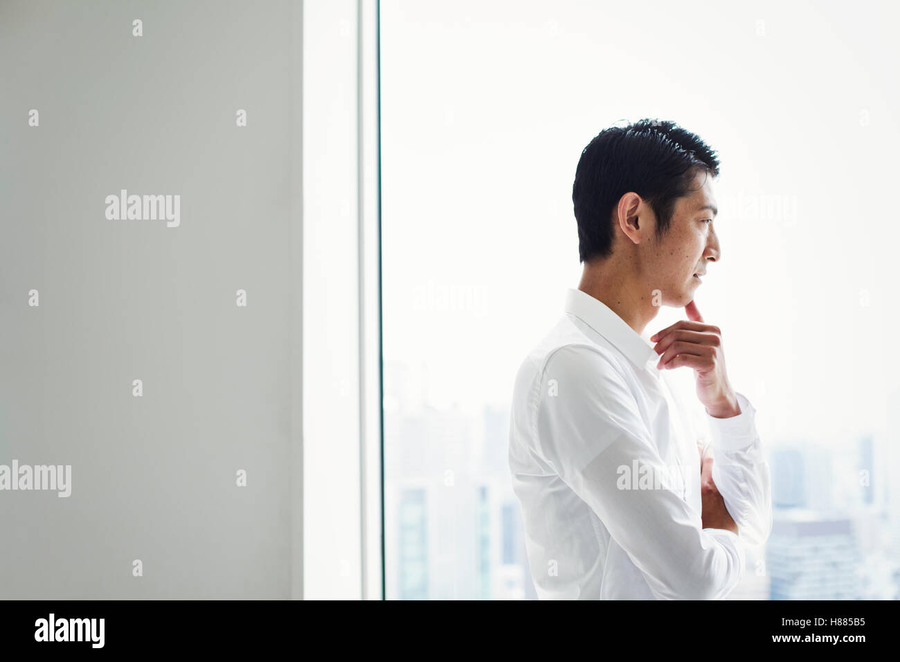 Un imprenditore in ufficio in piedi da una grande finestra con le braccia incrociate, guardando sopra la città. Foto Stock