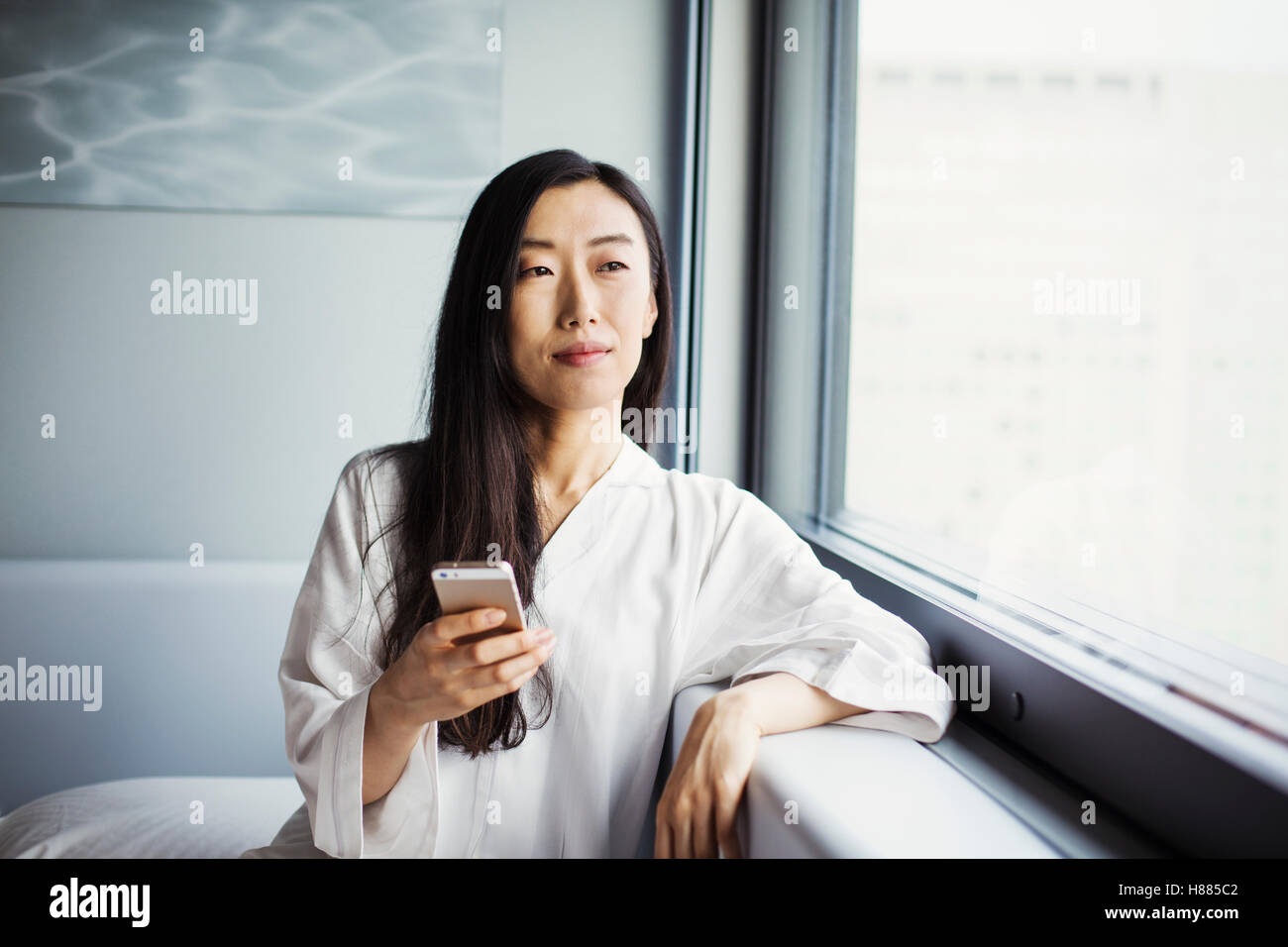 Una donna d'affari in preparazione per il lavoro, seduti in una finestra nel suo nightclothes, tenendo in mano un telefono intelligente. Foto Stock