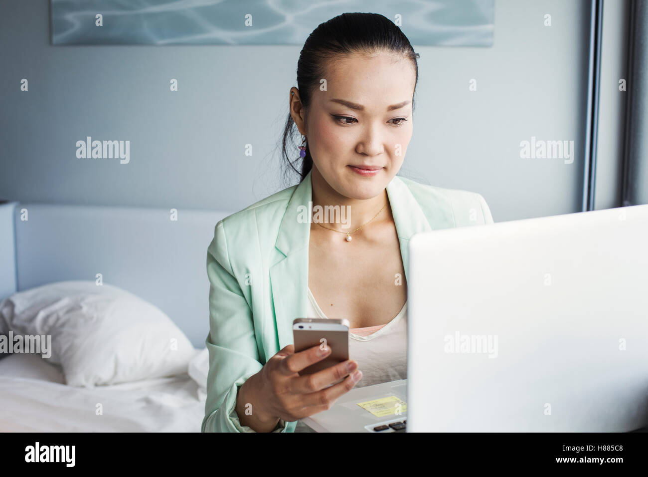 Una donna d'affari in preparazione per il lavoro, seduto su un letto mediante un computer portatile e tenendo il suo smart phone. Foto Stock