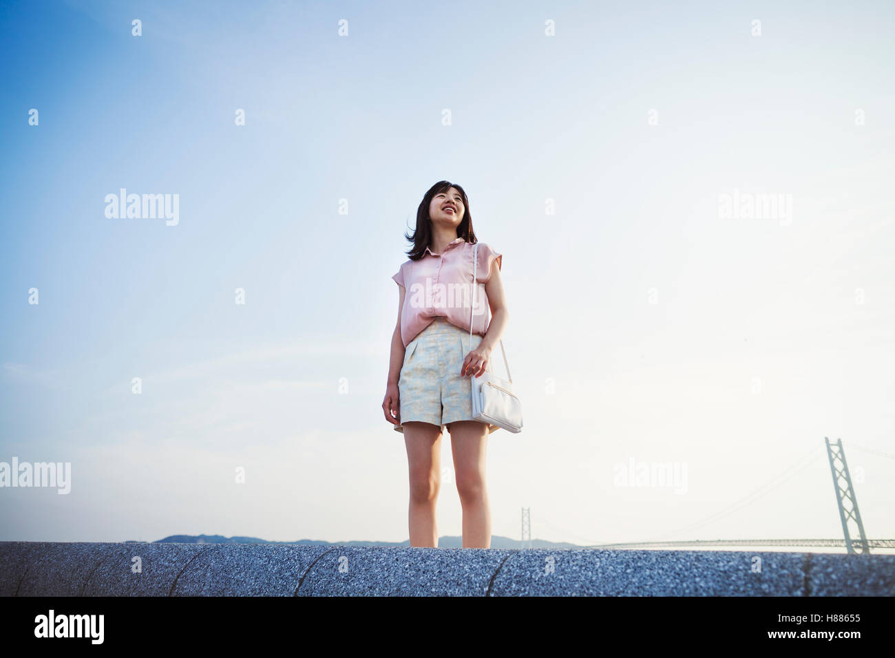 Giovane donna in pantaloncini e maglietta rosa solo in piedi in uno spazio aperto, con un grande ponte sospeso in background Foto Stock