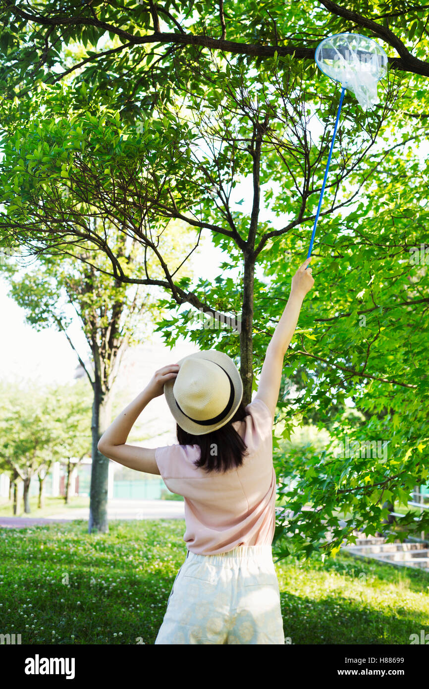 Vista posteriore di una giovane donna con capelli lunghi marrone in piedi in un parco, tenendo una farfalla net. Foto Stock