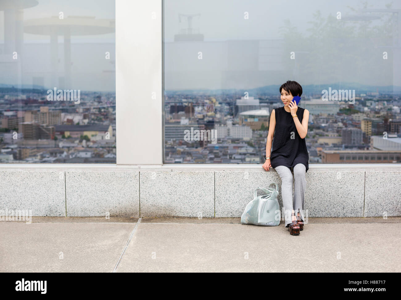 Una donna seduta da una finestra con vista su di una grande città con il suo ritorno alla vista, esecuzione di una chiamata su un telefono intelligente. Foto Stock