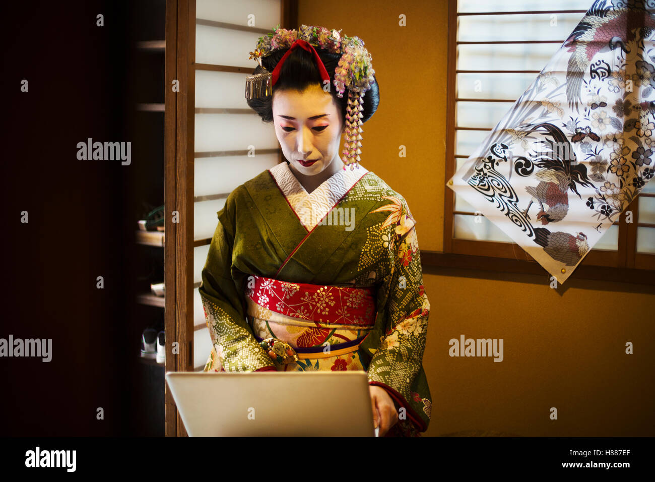 Una Donna vestita con il tradizionale stile geisha, indossando un kimono e OBI, con un elaborato e acconciatura di capelli floreali clip, con faccia bianca specchio luminoso con labbra rosse e occhi scuri usando un computer portatile. Foto Stock