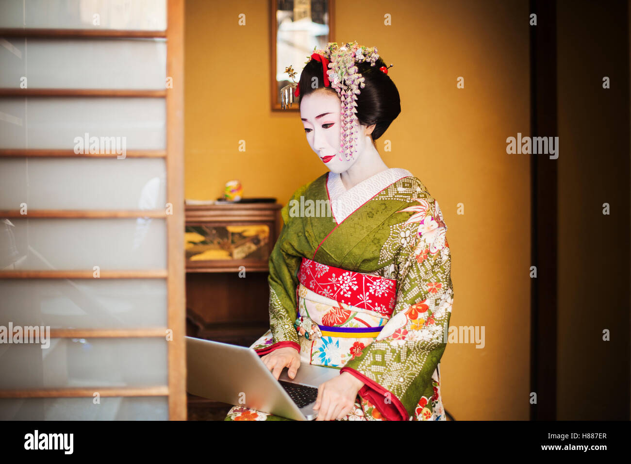 Una Donna vestita con il tradizionale stile geisha, indossando un kimono e OBI, con un elaborato e acconciatura di capelli floreali clip, con faccia bianca specchio luminoso con labbra rosse e occhi scuri usando un computer portatile. Foto Stock