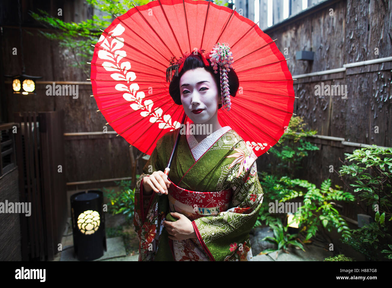 Una Donna vestita in stile geisha, bianco viso trucco con luminosi rossi labbra in possesso di una carta di colore rosso ombrellone. Foto Stock
