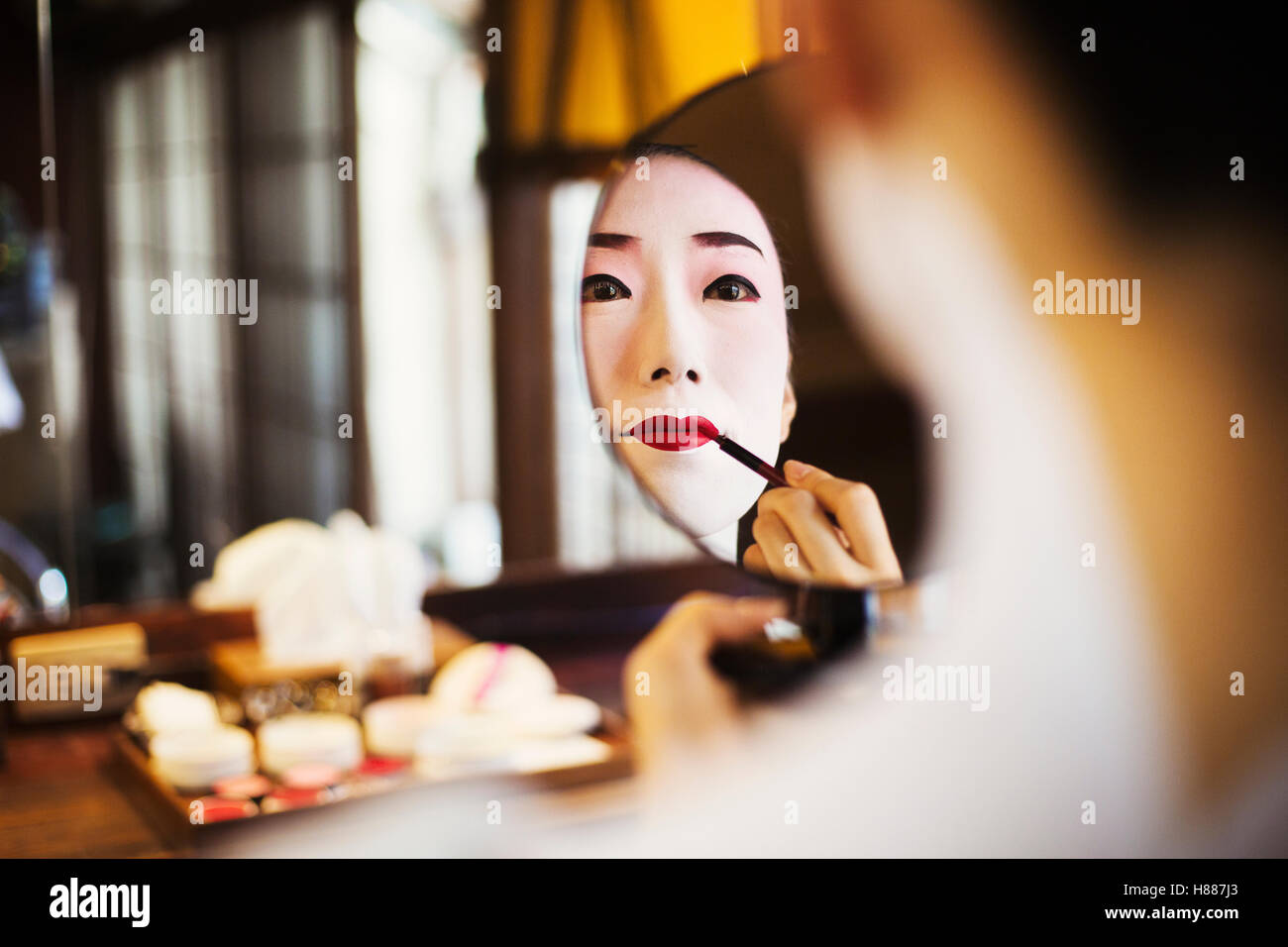 Geisha donna con bianco tradizionale viso trucco applicando rosso brillante rossetto con un pennello guardando in uno specchio. Foto Stock