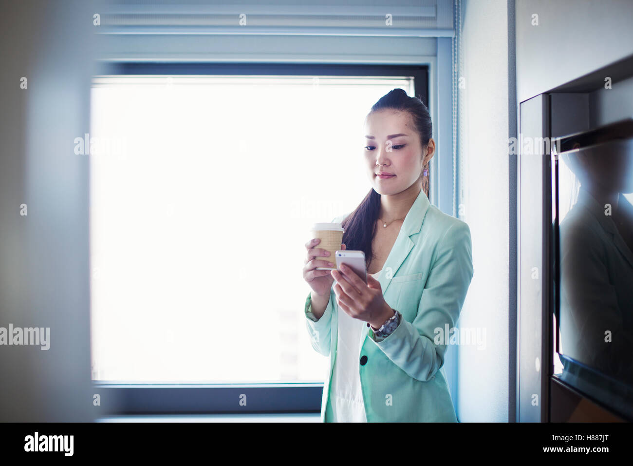 Una donna d'affari in preparazione per il lavoro, tenendo in mano una tazza di caffè e controllando il suo smart phone. Foto Stock