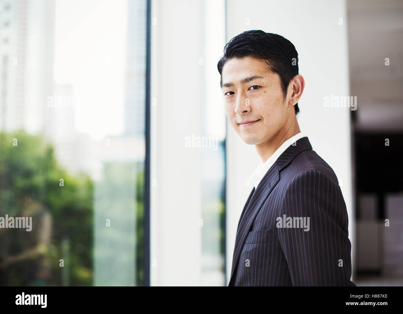 Un imprenditore in ufficio, da una grande finestra, guardando la telecamera. Indossare giacca e cravatta, vista sopra la città capo rovesciato Foto Stock