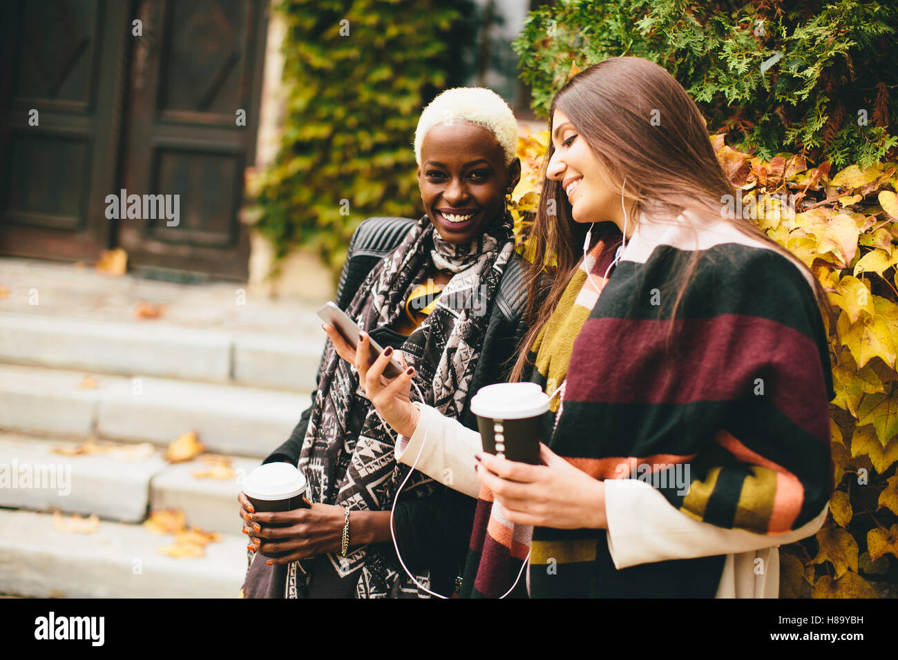 Americano africano e caucasici che pongono la donna fuori con il telefono cellulare e una tazza di caffè per andare in autunno Foto Stock