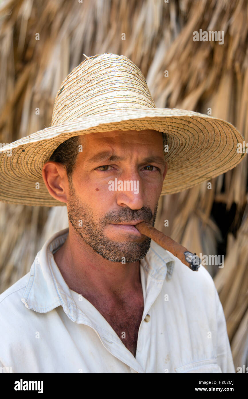 Ritratto di un giovane contadino cubano che indossa un cappello di paglia che fuma un sigaro con la casa di essiccazione del tabacco di paglia sullo sfondo a Vinales Cuba Foto Stock