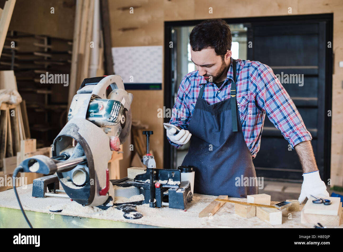 Carpenter texting qualcuno del suo smart phone vicino a sega circolare in un polveroso workshop Foto Stock