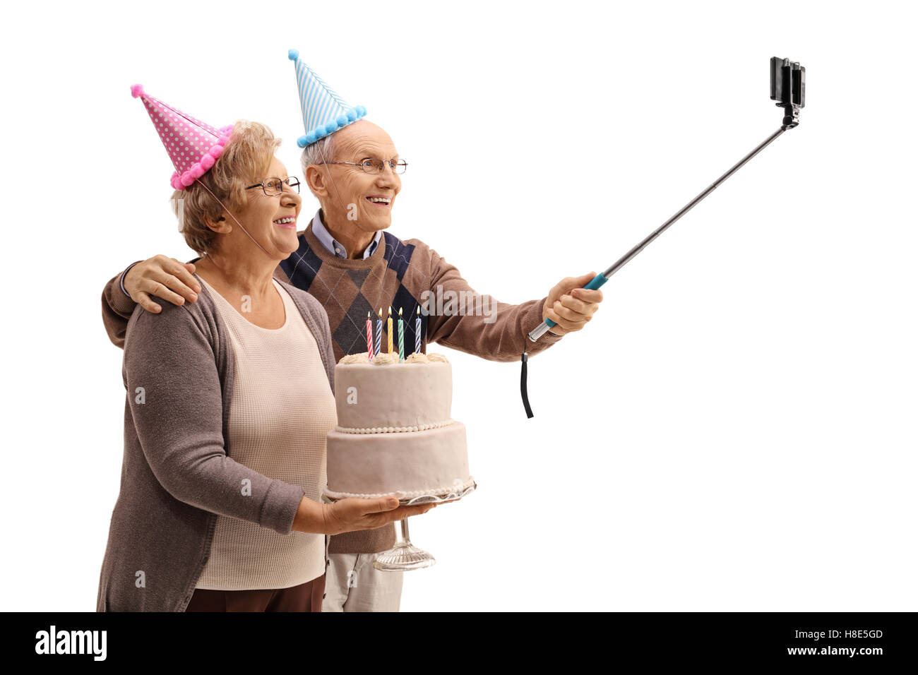 Felice di anziani con party cappelli e una torta di compleanno prendendo un selfie con un bastone isolato su sfondo bianco Foto Stock