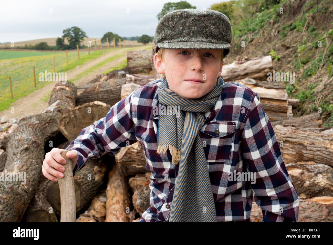 Ragazzo in plaid shirt, sciarpa e cappello tenendo un'ascia di fronte ad un mucchio di legna da ardere in una fattoria Foto Stock