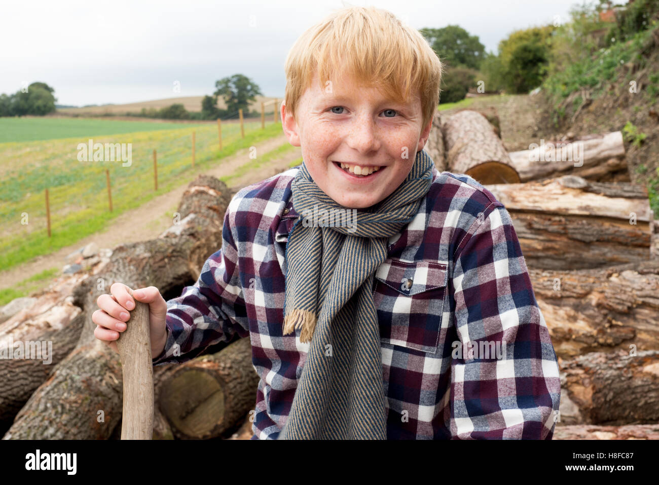 Ragazzo in plaid shirt, sciarpa e cappello tenendo un'ascia di fronte ad un mucchio di legna da ardere in una fattoria Foto Stock