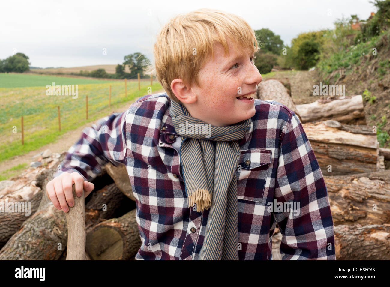 Ragazzo in plaid shirt, sciarpa e cappello tenendo un'ascia di fronte ad un mucchio di legna da ardere in una fattoria Foto Stock