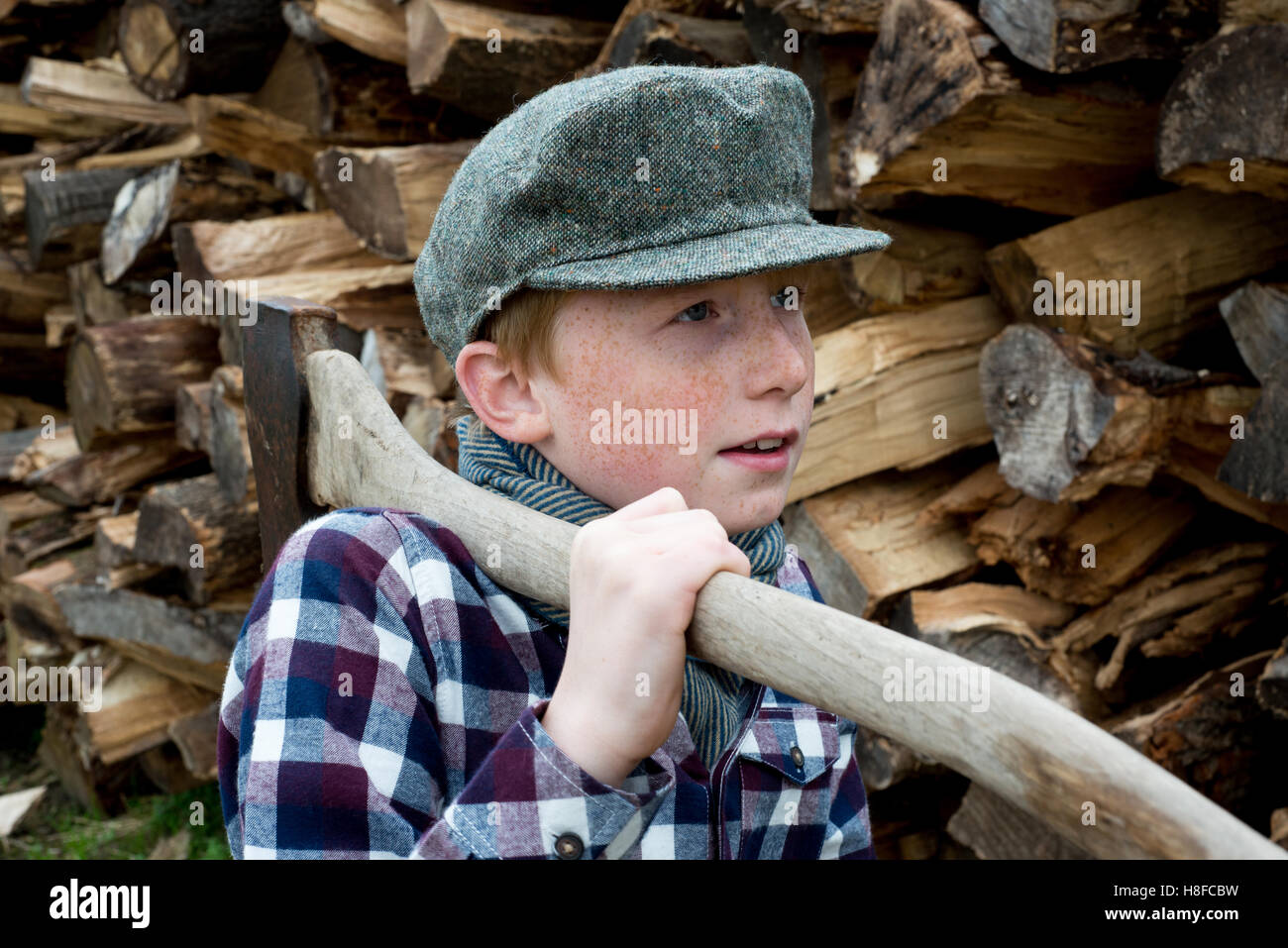 Ragazzo in plaid shirt, sciarpa e cappello tenendo un'ascia di fronte ad un mucchio di legna da ardere in una fattoria Foto Stock