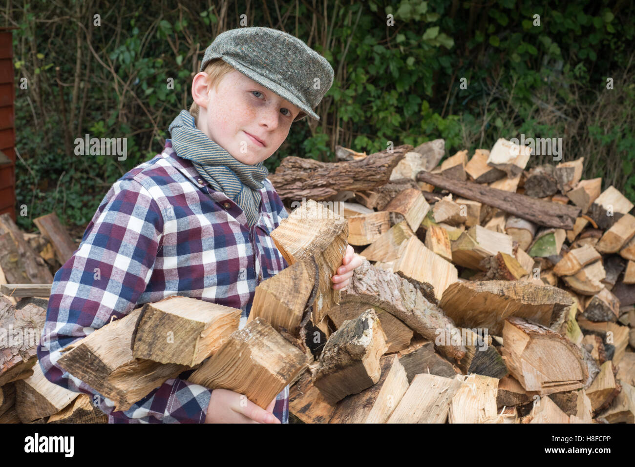 Ragazzo in plaid shirt, sciarpa e cappello tenendo un'ascia di fronte ad un mucchio di legna da ardere in una fattoria Foto Stock