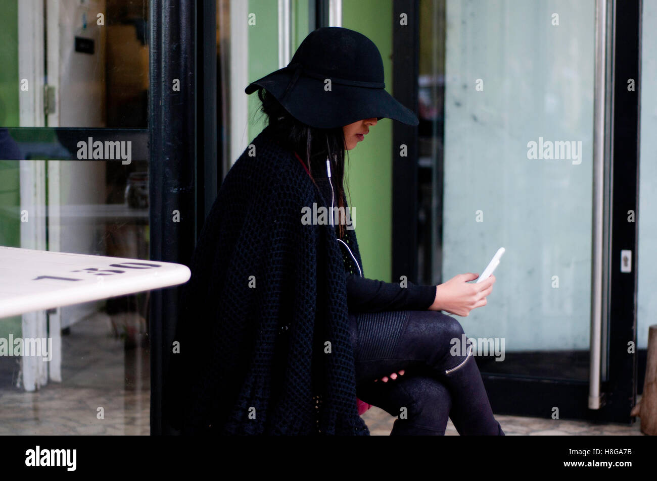 Giovane donna asiatica con big black hat, usando il cellulare mentre è seduto a Londra. Foto Stock