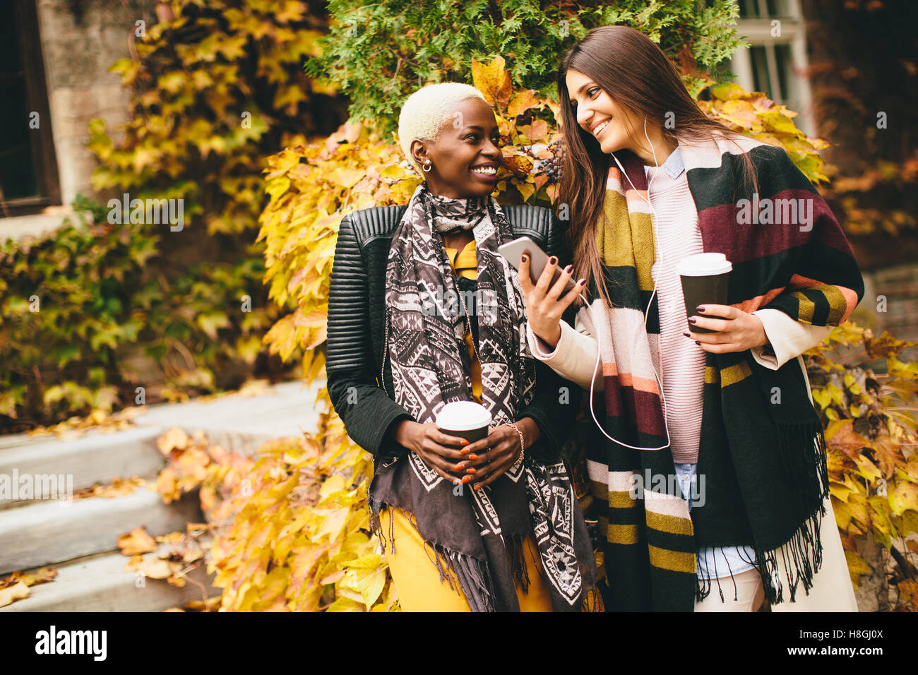 Americano africano e caucasici che pongono la donna fuori con il telefono cellulare e una tazza di caffè per andare in autunno Foto Stock