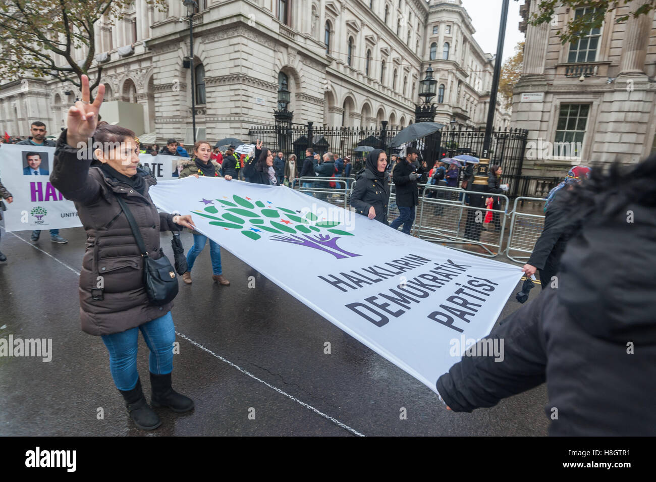 Londra, Regno Unito. 12 novembre 2016. Una donna con il banner per l opposizione Partito Democratico del Popolo (HDP) almeno 9 di cui MPs inclusi i due leader sono stati arrestati fa un v per la vittoria di segni come andare passato Downing St. centinaia di curdi e turchi marzo a Londra per difendere la pace e la democrazia in Turchia contro le azioni del Presidente Erdogan. Poiché i militari flebile tentativo di colpo di stato di luglio, Erdogan e il suo governo AKP ha imposto uno stato di emergenza, chiusura 170 media outlet, l'arresto 128 giornalisti, spurgando almeno 110.000 lavoratori del settore pubblico inclusi 11.000 Foto Stock