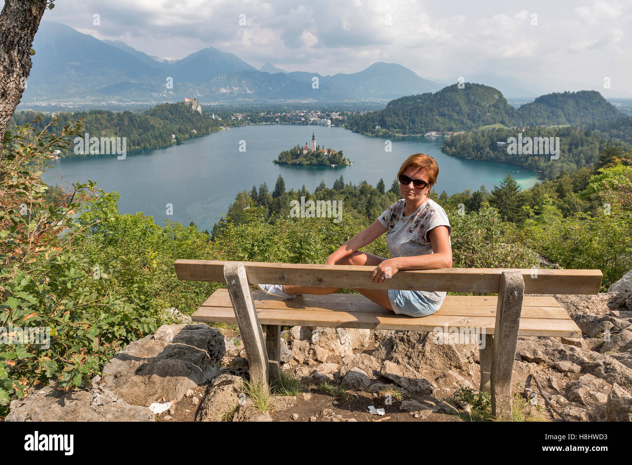 Bianco di mezza età donna abbronzata sedersi sulla panchina di legno con vista dal di sopra del lago di Bled. Dalla montagna Osojnica in Slovenia. Foto Stock
