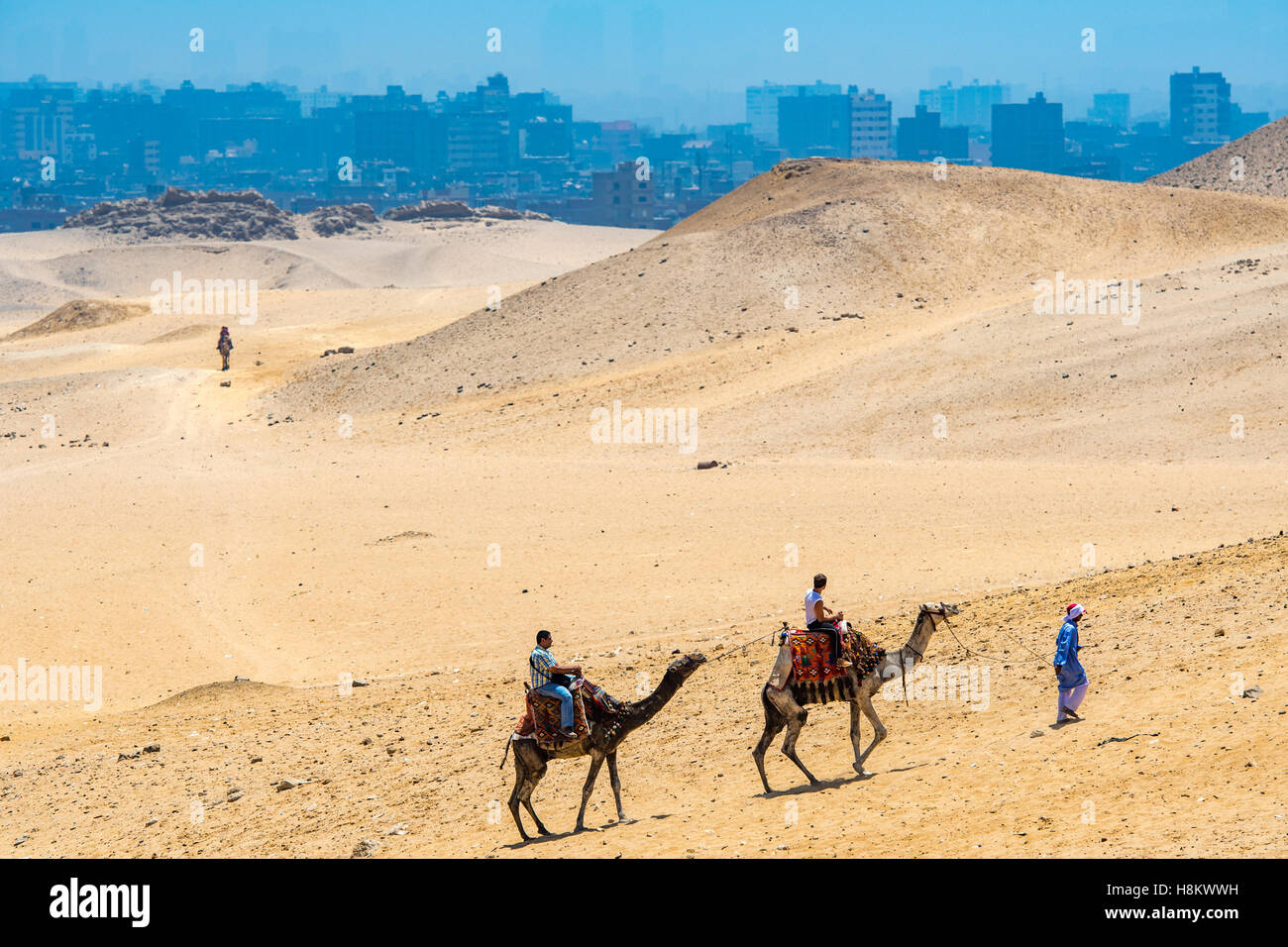 Cairo, Egitto driver del cammello e turisti a cavallo di cammelli a piedi attraverso il deserto con la città del Cairo in background. Foto Stock