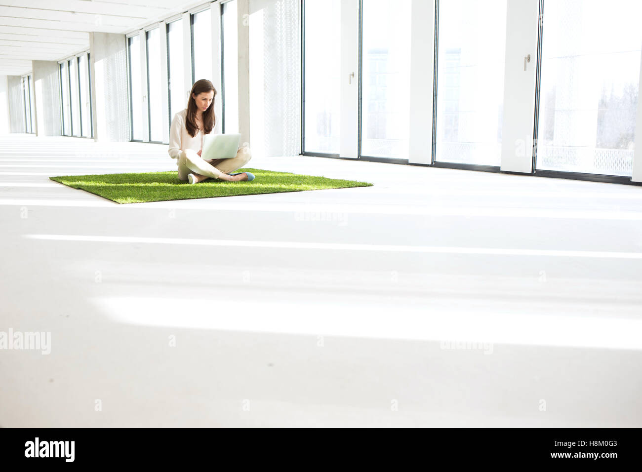Giovane imprenditrice utilizzando laptop stando seduti sul manto erboso in ufficio vuoto Foto Stock
