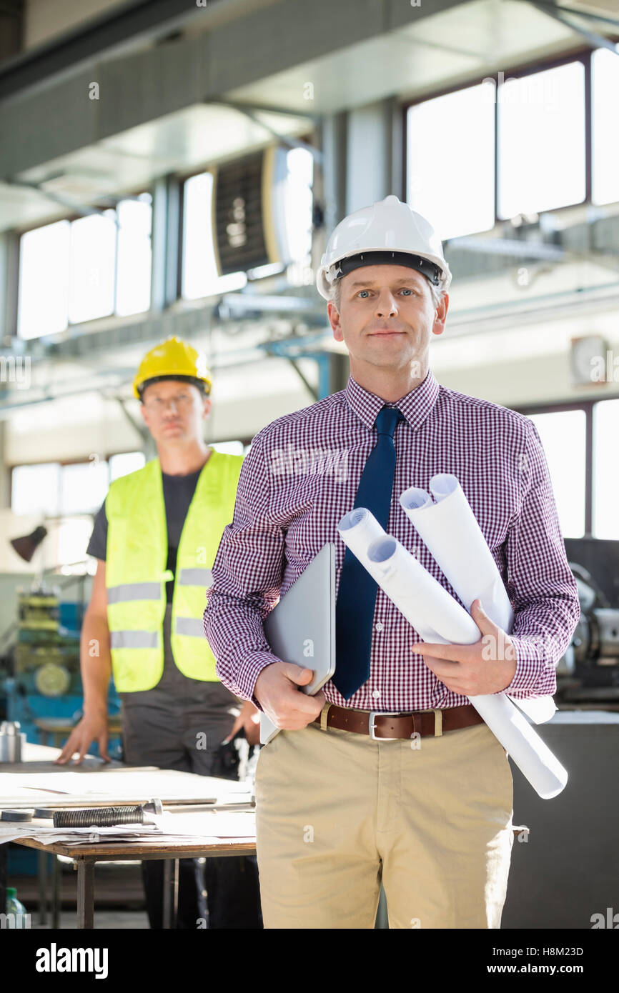 Ritratto di fiducioso architetto maschio holding blueprint e portatile con il lavoratore in background all'industria Foto Stock