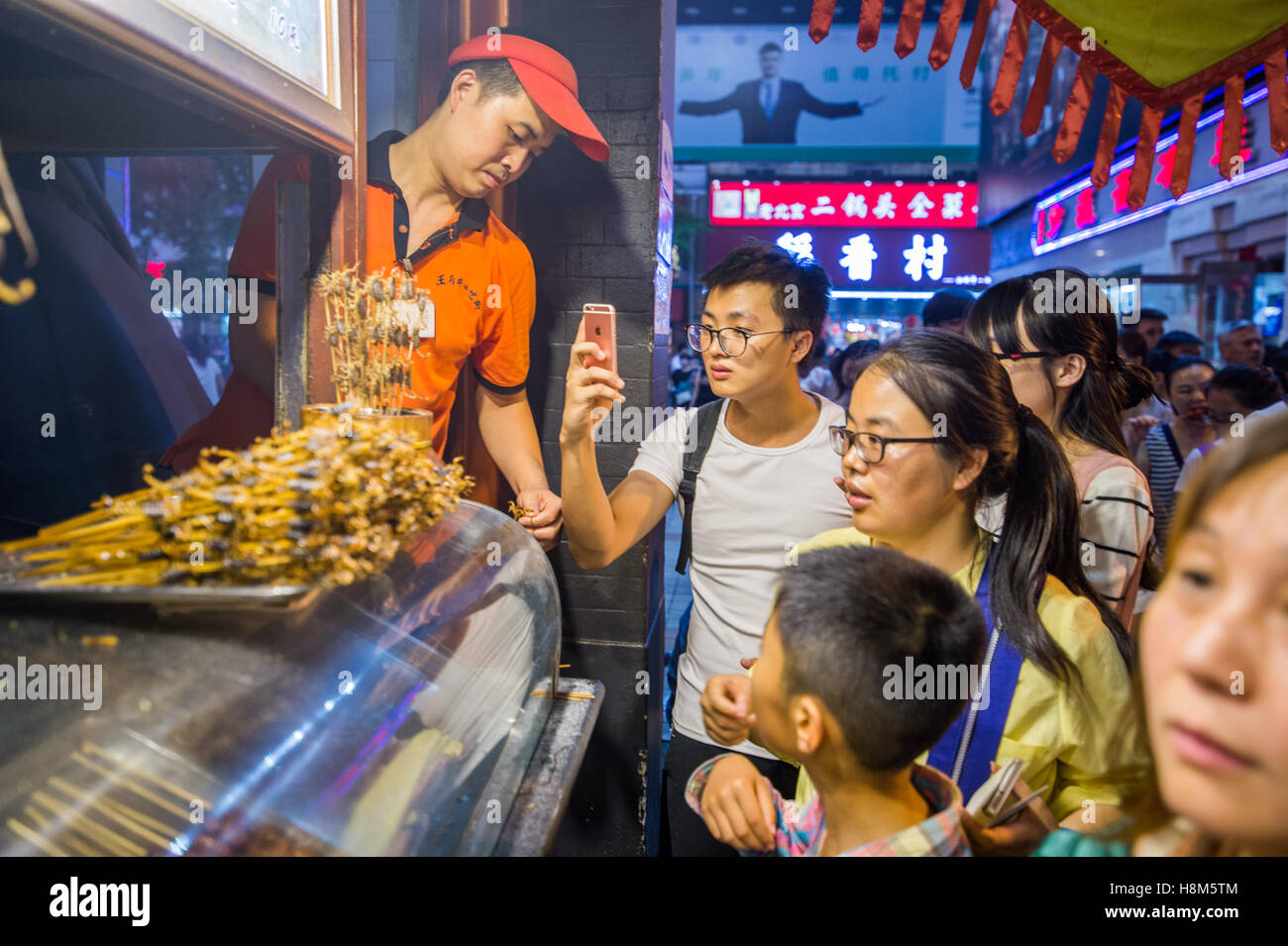 Pechino, Cina - Maschio turistica prendendo una foto sul suo telefono delle cellule di scorpioni su bastoni in vendita presso la Donghuamen Snack notte M Foto Stock