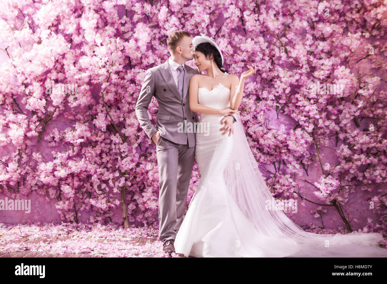 Romantico sposo baciare sposa sulla fronte mentre in piedi contro la parete ricoperta con fiori di colore rosa Foto Stock