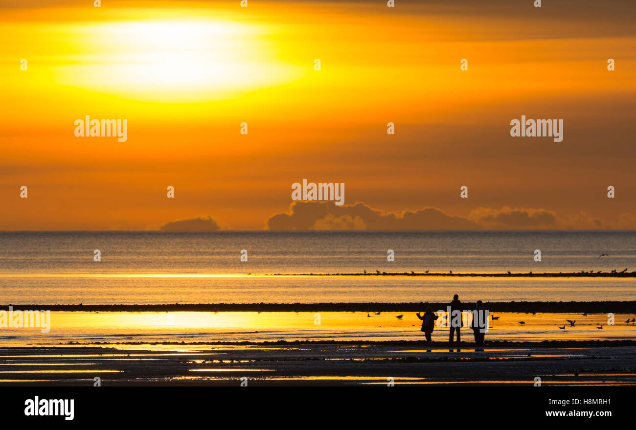 Impostazione di sole in un giorno nuvoloso lungo la costa con una piccola famiglia in silhouette guardando. Foto Stock