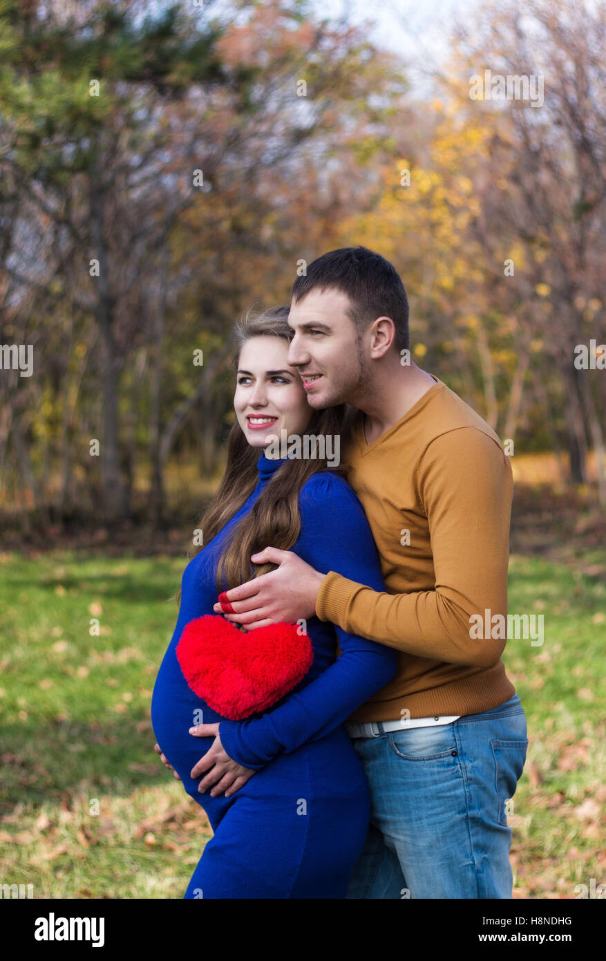 Giovane coppia incinta in piedi nel parco. Uomo che abbraccia una donna da dietro e detiene un cuore morbido forma toy .stagione autunnale. Foto Stock