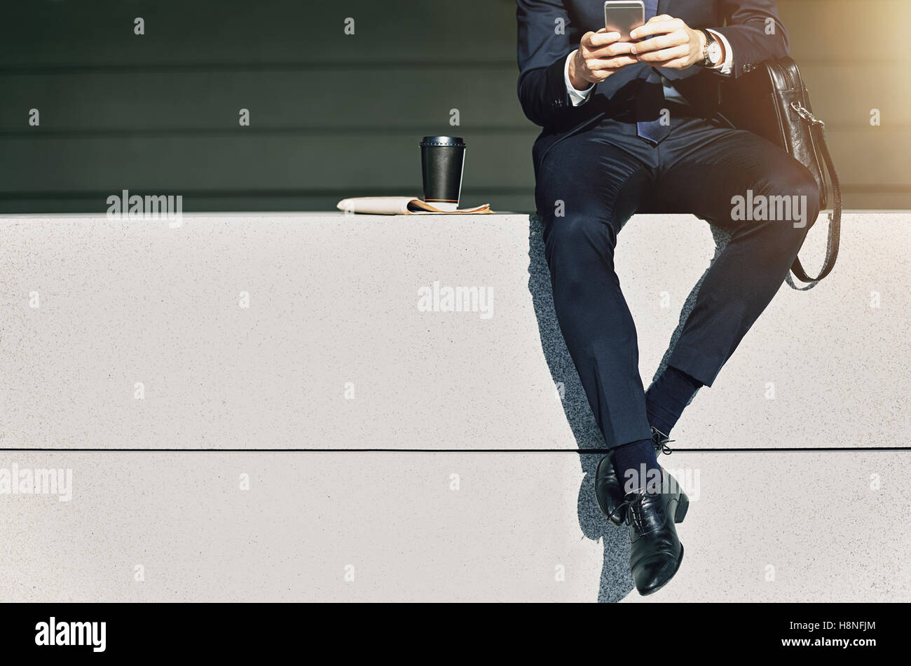 Maschio di raccolto di indossare tuta seduto sul parapetto con tazza di caffè vicino e utilizzando il telefono Foto Stock