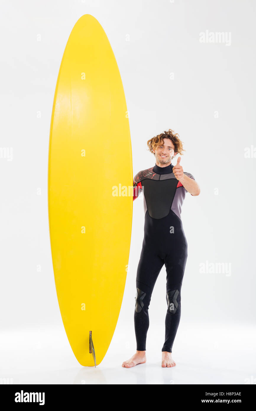 Felice giovani surfer tenendo la tavola da surf e mostrando i pollici fino gesto isolato su sfondo bianco Foto Stock