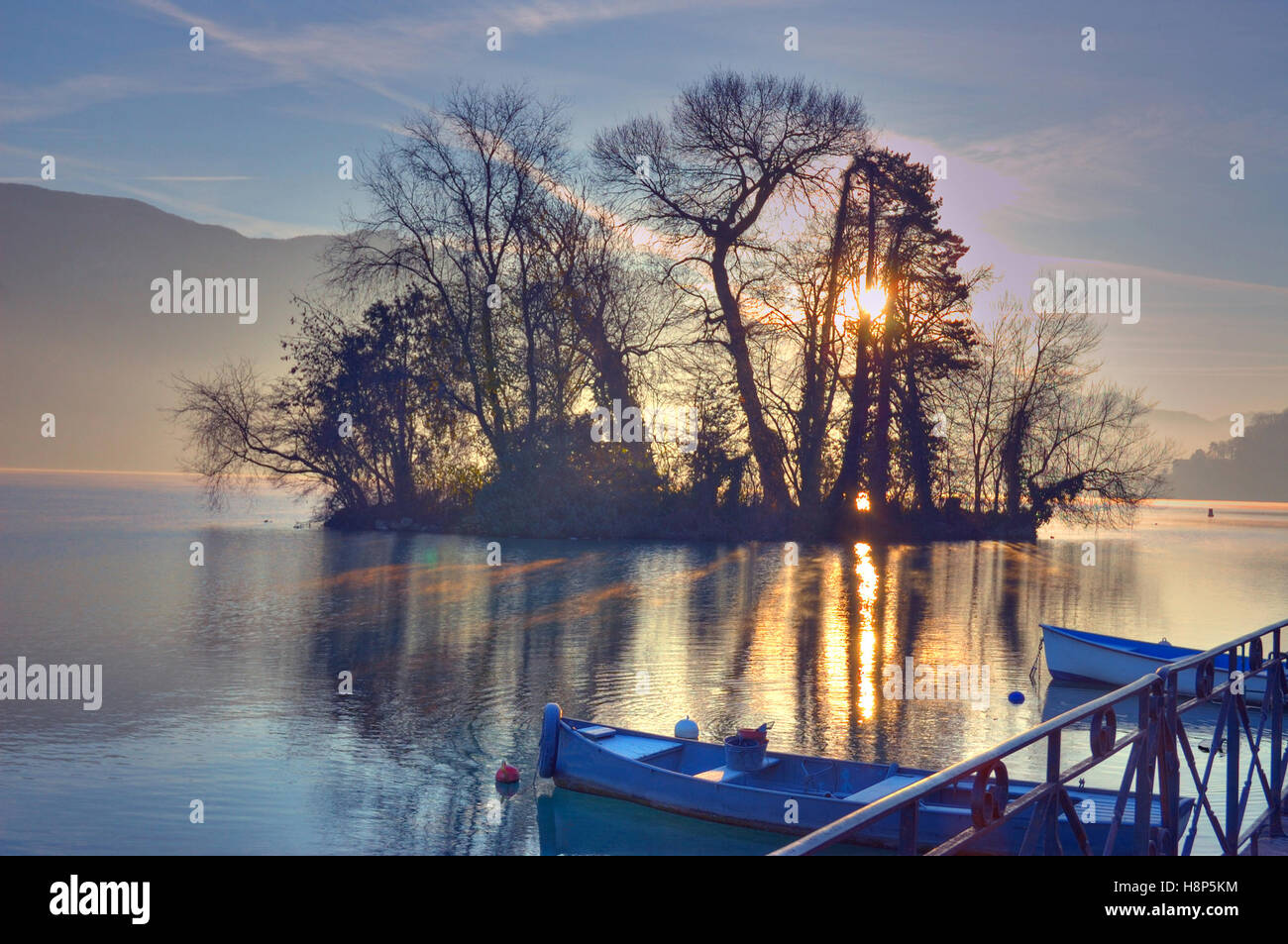 La luce del mattino attraverso gli alberi su una piccola isola nel lago di Annecy Francia con piccole barche da pesca legato in primo piano. Foto Stock