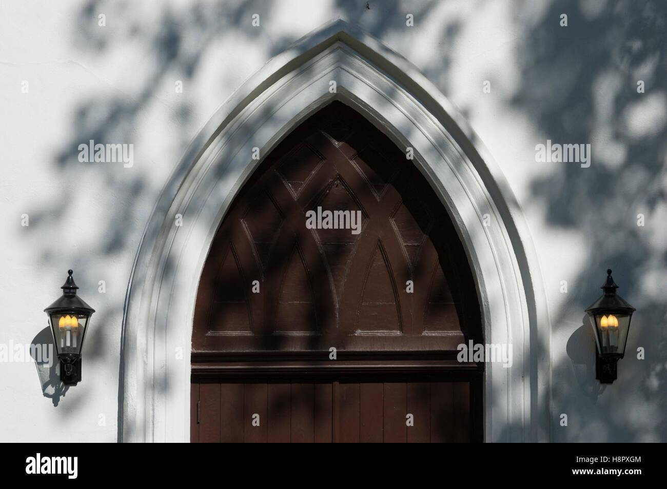 Un arcuato marrone vecchia porta della chiesa fiancheggiata da lampade e cosparsi di ombre di un albero. Foto Stock