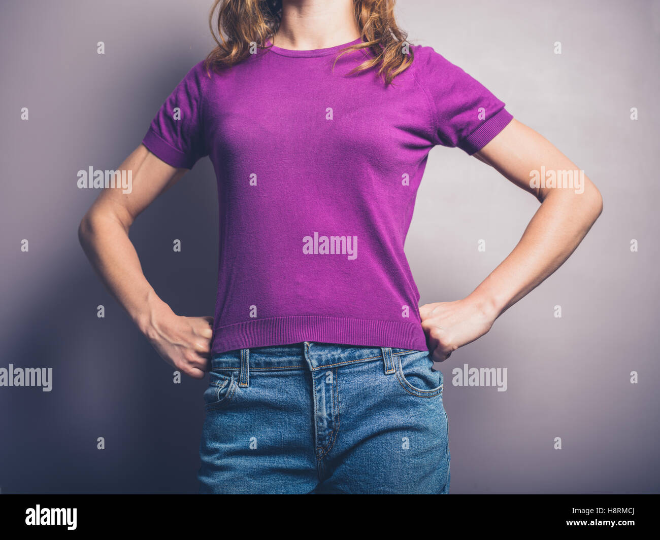 Un fiducioso giovane donna in maglia viola è in piedi in un potente posano con la sua mani sui fianchi Foto Stock