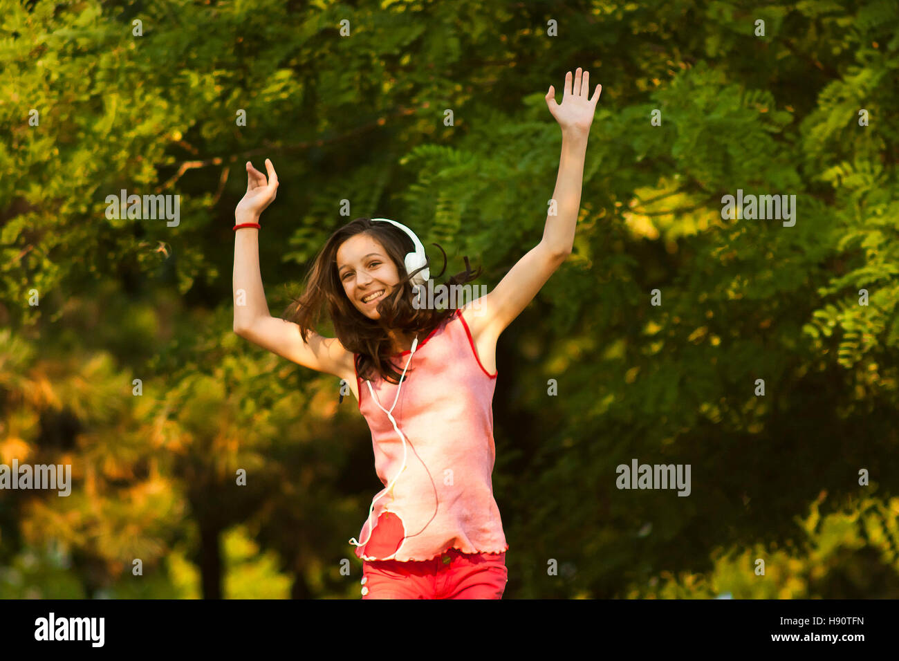 Felice ragazza adolescente in rosso dancing in natura mentre si ascolta la musica con le cuffie, sorridente e guardando la telecamera su un soleggiato giorno d'estate. Foto Stock