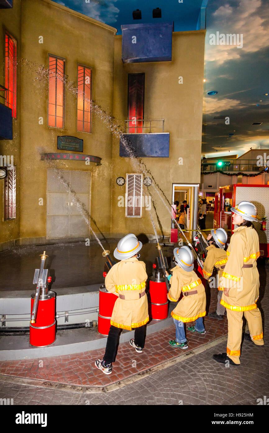 Giappone, Nishinomiya, KidZania, attività figlio il centro del gioco. Linea di bambini vestiti come i vigili del fuoco mirando tubi flessibili a combustione edificio. Foto Stock