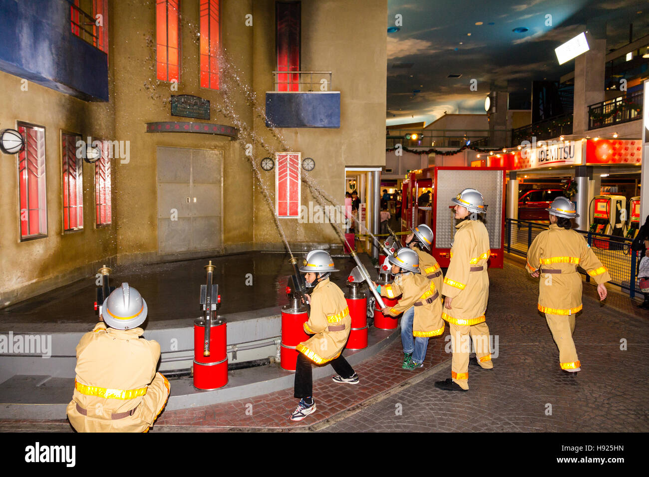 Giappone, Nishinomiya, KidZania, attività figlio il centro del gioco. Linea di bambini vestiti come i vigili del fuoco mirando tubi flessibili a combustione edificio. Foto Stock