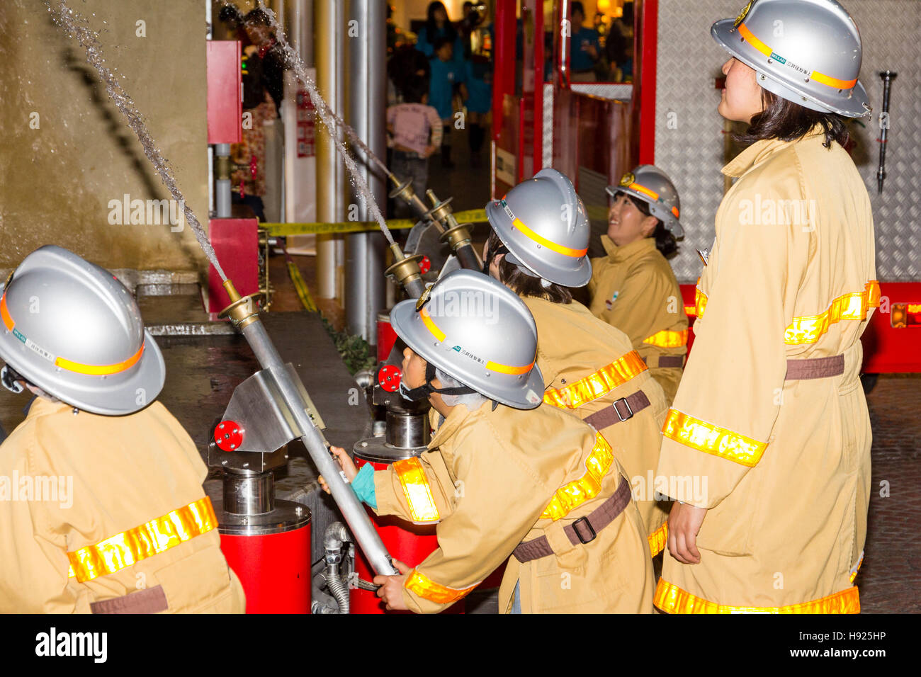 Giappone, Nishinomiya, KidZania, attività figlio il centro del gioco. Linea di bambini vestiti come i vigili del fuoco mirando tubi flessibili a combustione edificio. Foto Stock